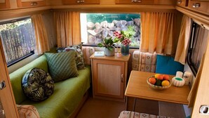 Een gezellig interieur van een camper, met een groene bank, een kleine tafel met fruit en een raam met uitzicht op rotsen en groen.