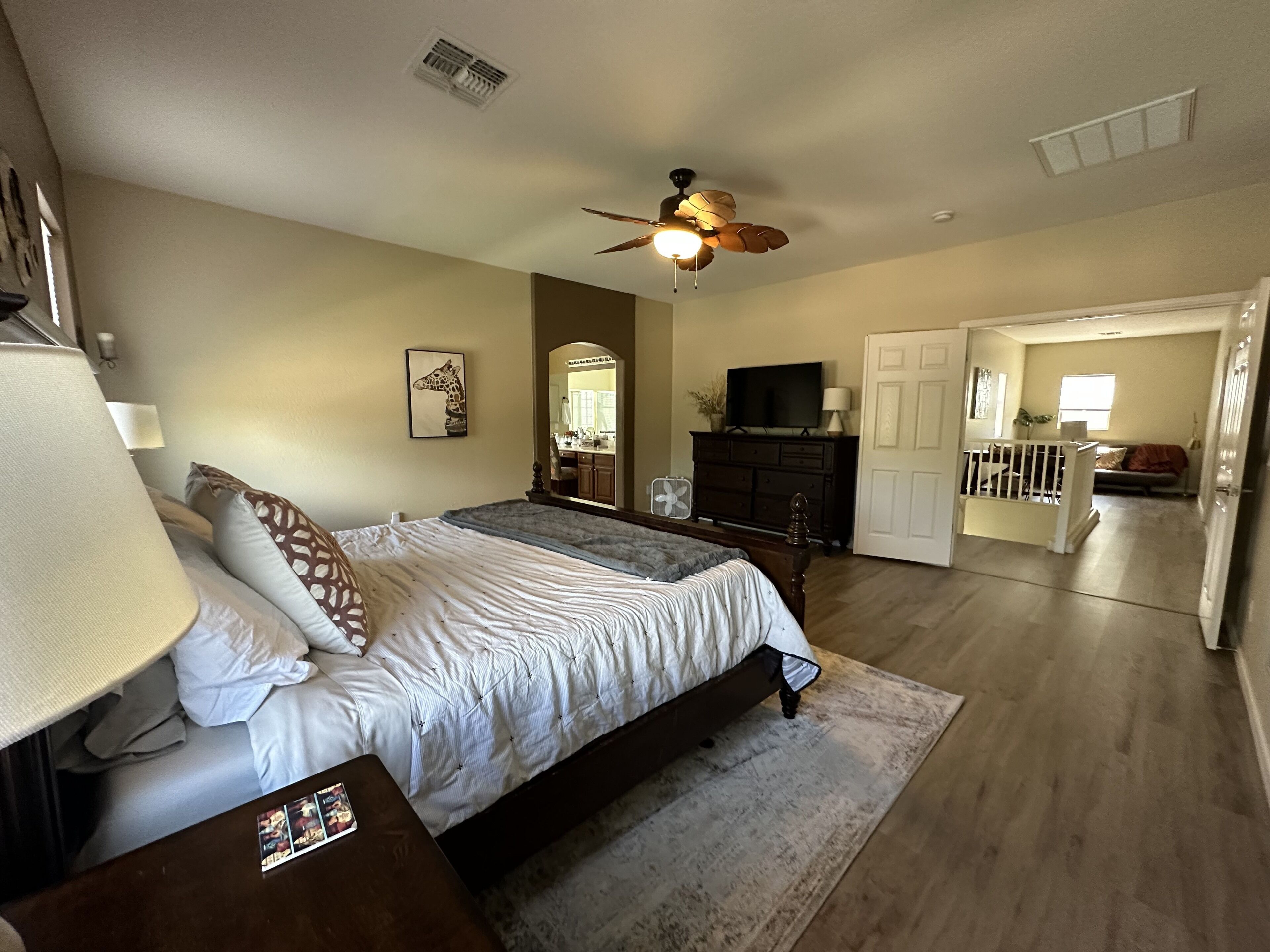 A bedroom with a bed, a ceiling fan, a dresser, a TV, and a door leading to another room.