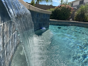 A clear view of a swimming pool with a waterfall feature, surrounded by a stone wall and lush greenery.