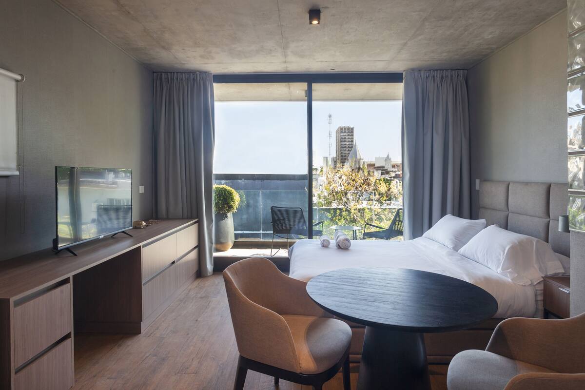 Una habitación de hotel moderna con una cama grande, un escritorio con televisión, un balcón con vista a la ciudad y un comedor con mesa y sillas.