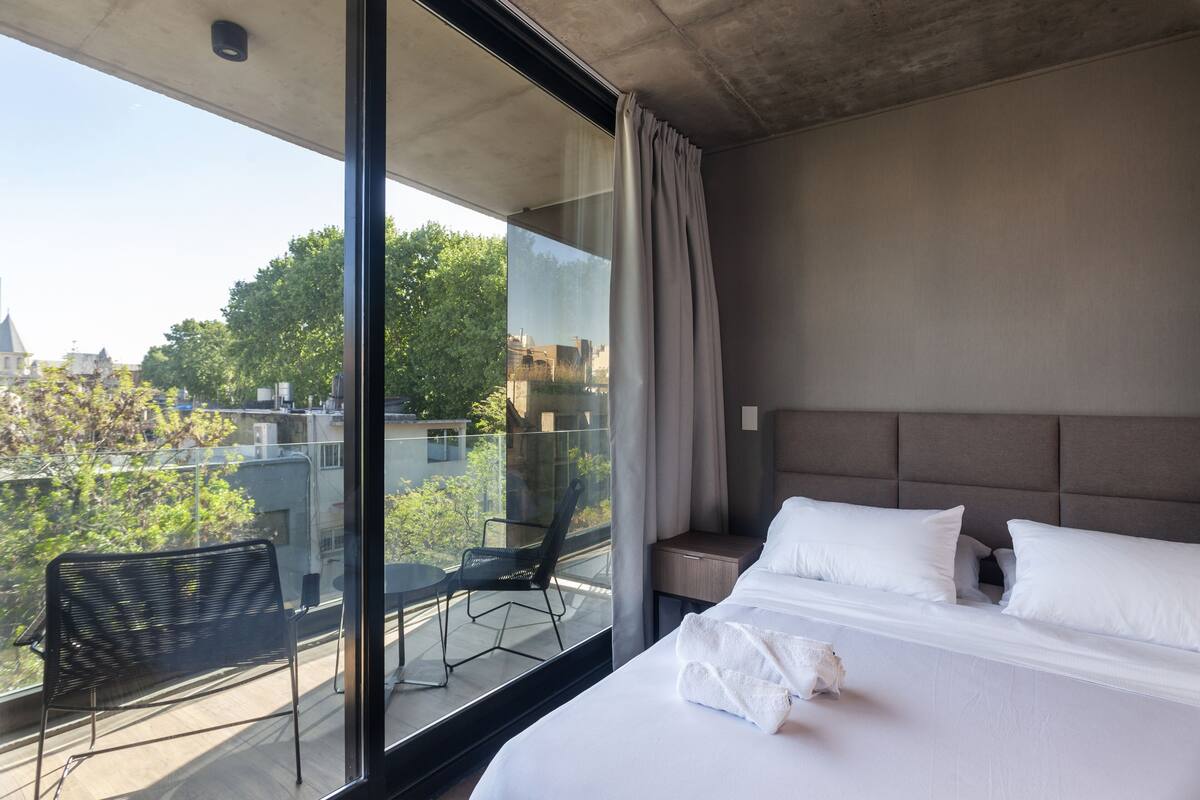 Habitación de hotel con una cama grande, vista de la ciudad desde un balcón y una silla afuera.
