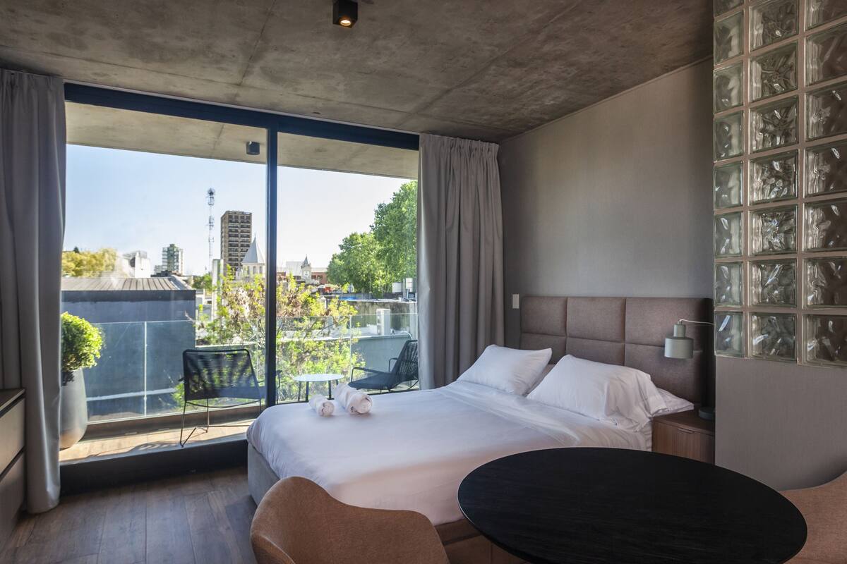 Una habitación de hotel moderna con una cama grande, una mesa redonda y un balcón con vistas a la ciudad.