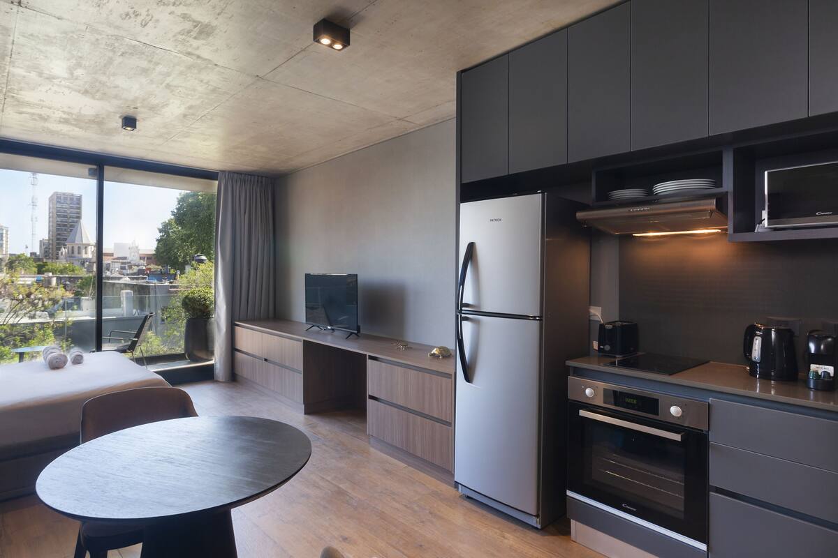 Una cocina moderna con refrigerador, horno y microondas incorporados. Hay un comedor con mesa y sillas, y un amplio ventanal con vistas a la ciudad.