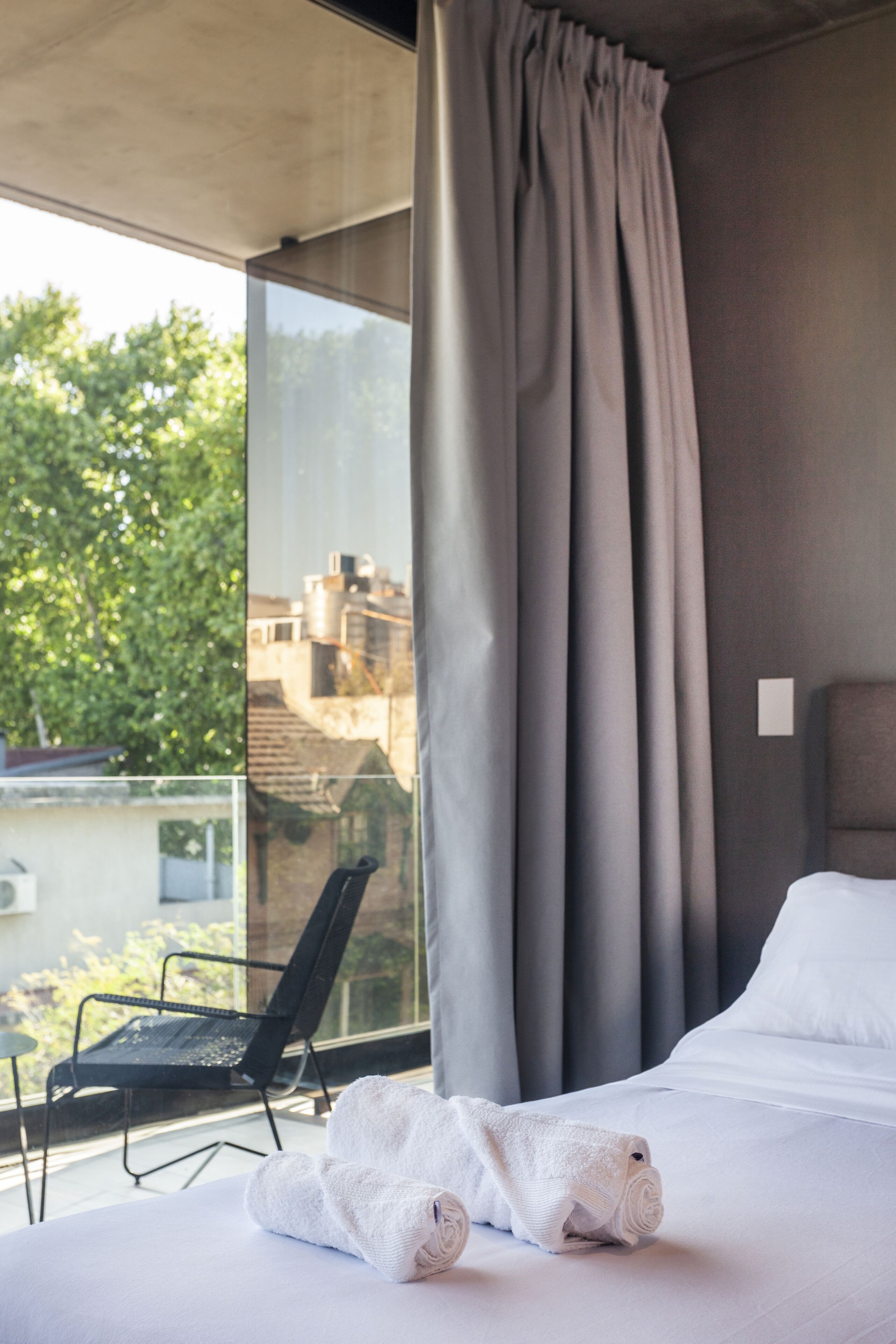 A hotel room with a bed, a window with curtains, a chair, and a view of buildings and trees outside.