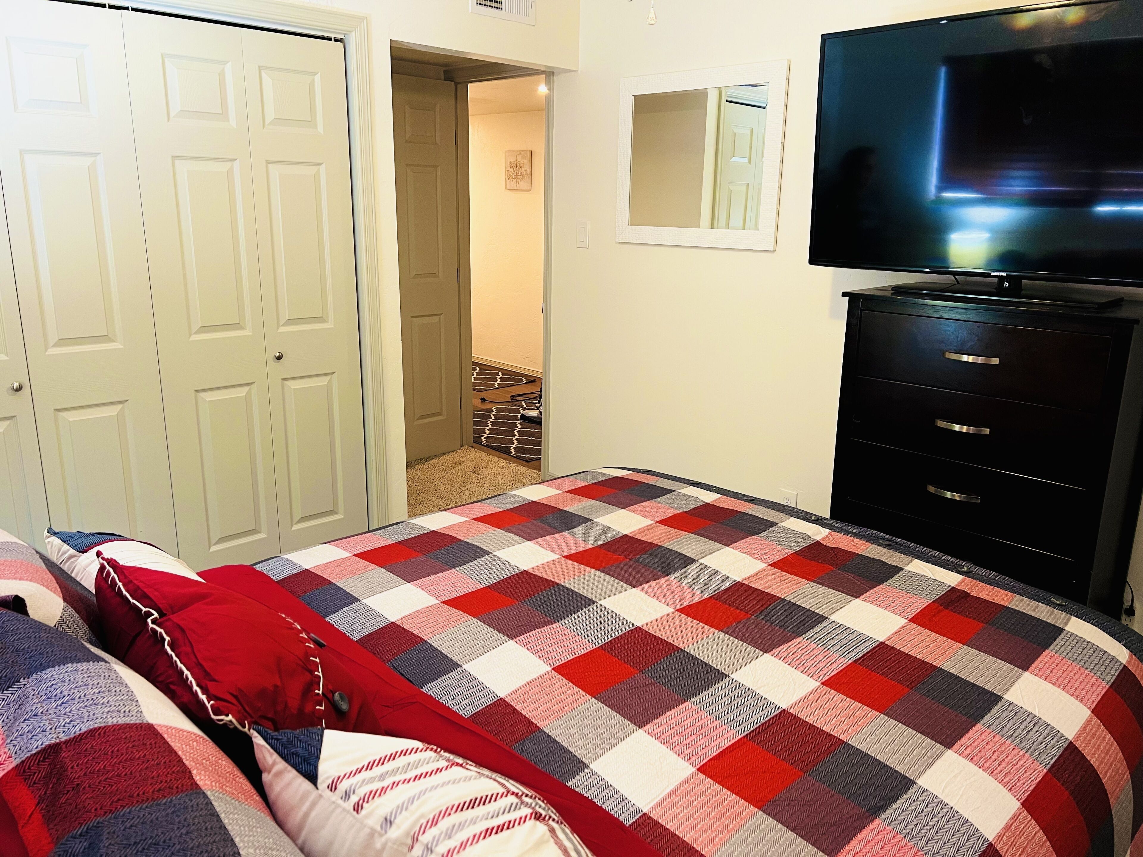 Een slaapkamer met een geruit beddegoed, een televisie, een dressoir en een deur die naar een gang leidt.