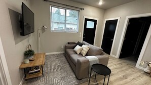 A compact living space with a sofa, a small table, a TV mounted on the wall, a window, and a door. - New Guest Suite 2br/1bth in DT Vancouver - pet friendly (Vancouver)