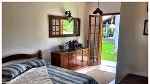 A bedroom with a bed, a wooden dresser, a mirror, and a door leading to a garden.