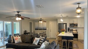 A modern living room with a dark-colored sofa, a dining area with a table and chairs, and a kitchen with white cabinets and stainless steel appliances.