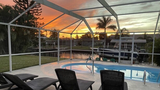 A covered outdoor area with a pool, lounge chairs, and a view of a sunset over water.