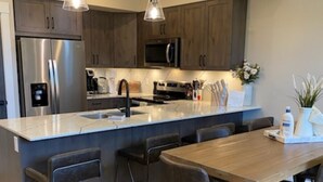 Een moderne keuken met roestvrijstalen apparatuur, een houten kookeiland met barstoelen, hanglampen en uitzicht op de woonkamer.