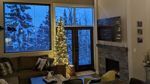 Een gezellige woonkamer met een open haard, een kerstboom en een groot raam met uitzicht op een besneeerd landschap.