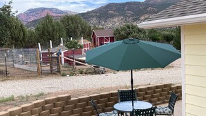 Une terrasse avec une table et des chaises, un parasol vert, et une vue sur les montagnes, ainsi qu’une zone délimitée par une clôture où se trouve une grange rouge.
