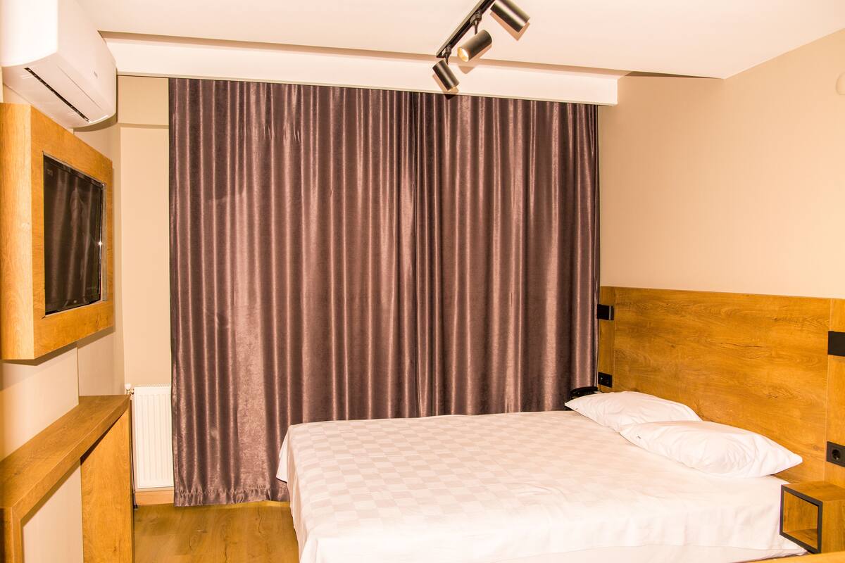 A hotel room with a wooden headboard, a bed with white linens, a large window with a view, and a wall-mounted air conditioner.