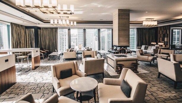 Un moderno vestíbulo de hotel con una zona de estar central, un mostrador de recepción y grandes ventanales con vistas a la ciudad.
