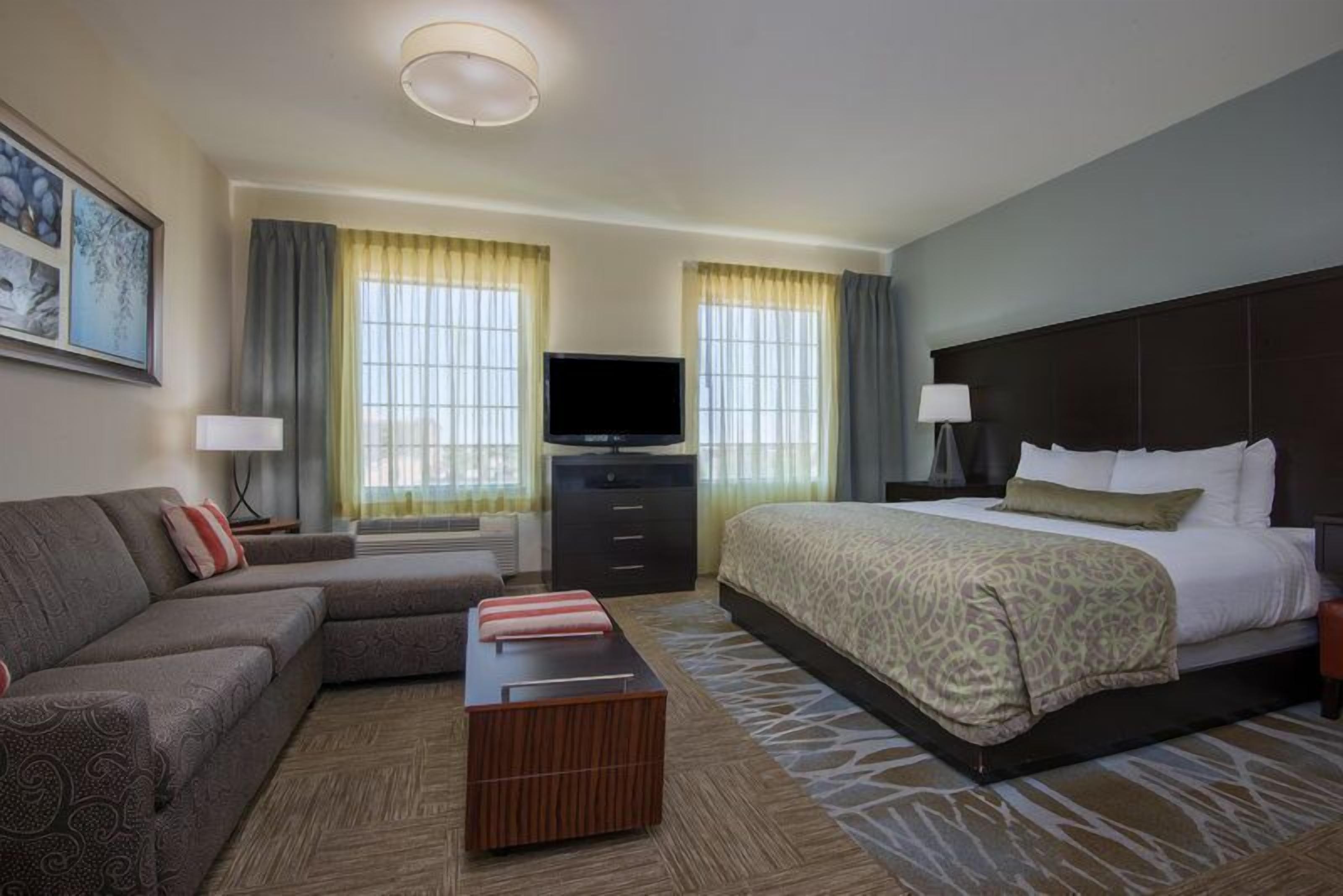 A hotel room with a bed, a sofa, a TV, a desk, and two windows with curtains.
