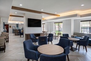 Un hall d’hôtel moderne avec des tables rondes, des chaises bleues, un téléviseur à écran plat et un coin bar.