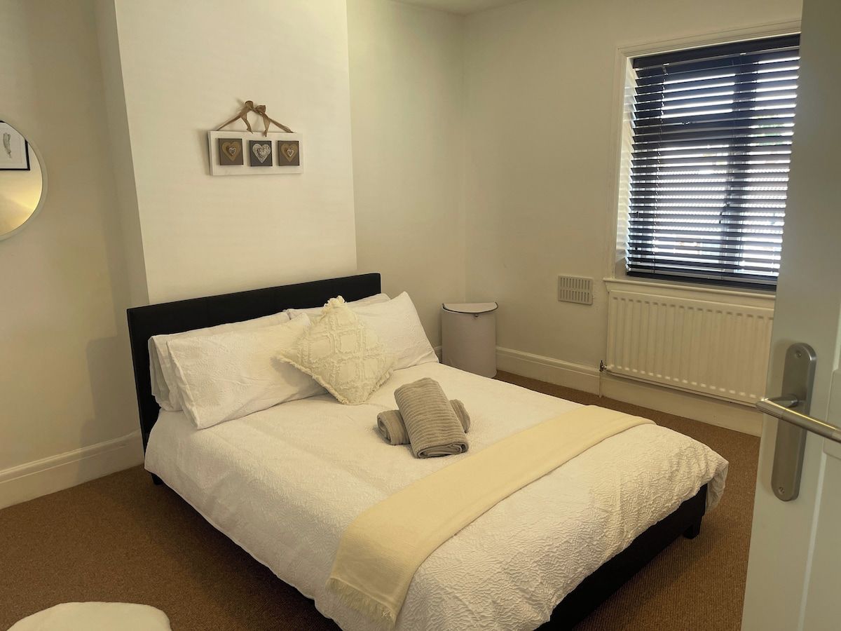 Ein ordentlich bezogenes Bett mit weißer Daunendecke und beige farbenem Handtuch, ein Nachttisch, ein an der Wand befestigter Bilderrahmen, ein Fenster mit Jalousien und ein Heizkörper.