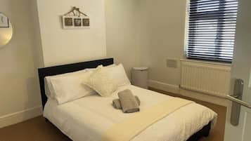 Ein ordentlich bezogenes Bett mit weißer Daunendecke und beige farbenem Handtuch, ein Nachttisch, ein an der Wand befestigter Bilderrahmen, ein Fenster mit Jalousien und ein Heizkörper.