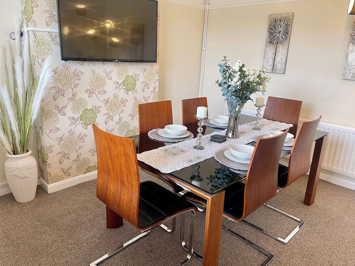 A dining area with a table set for four, surrounded by chairs, a vase with flowers, and a television mounted on the wall.