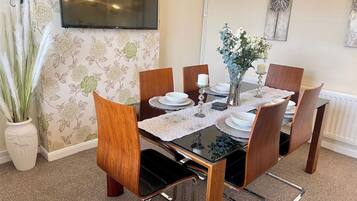 A dining area with a table set for four, surrounded by chairs, a vase with flowers, and a television mounted on the wall.