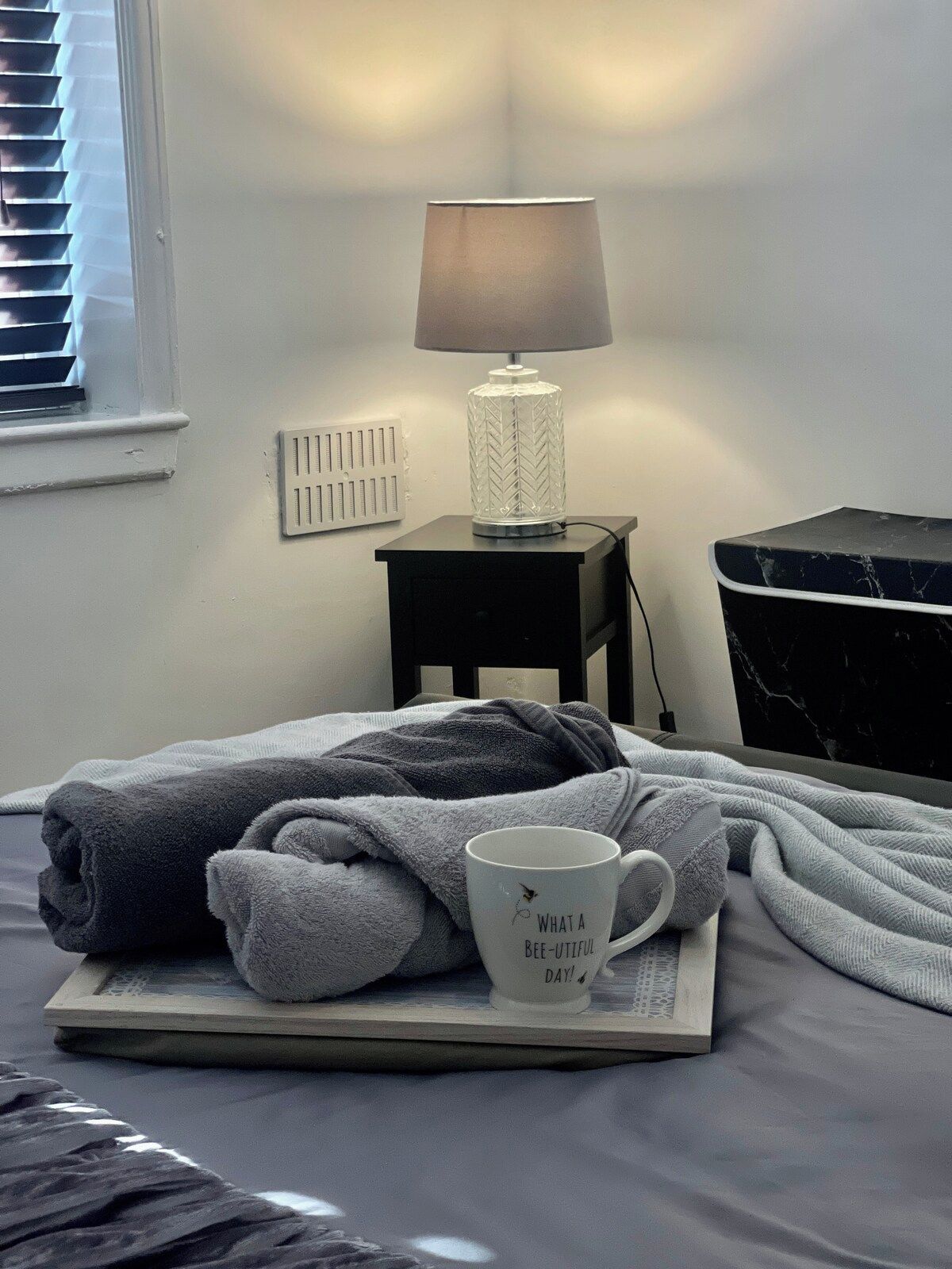 Una cama bien hecha con un edredón gris y una taza blanca con texto, una mesita de noche con lámpara y un armario negro.