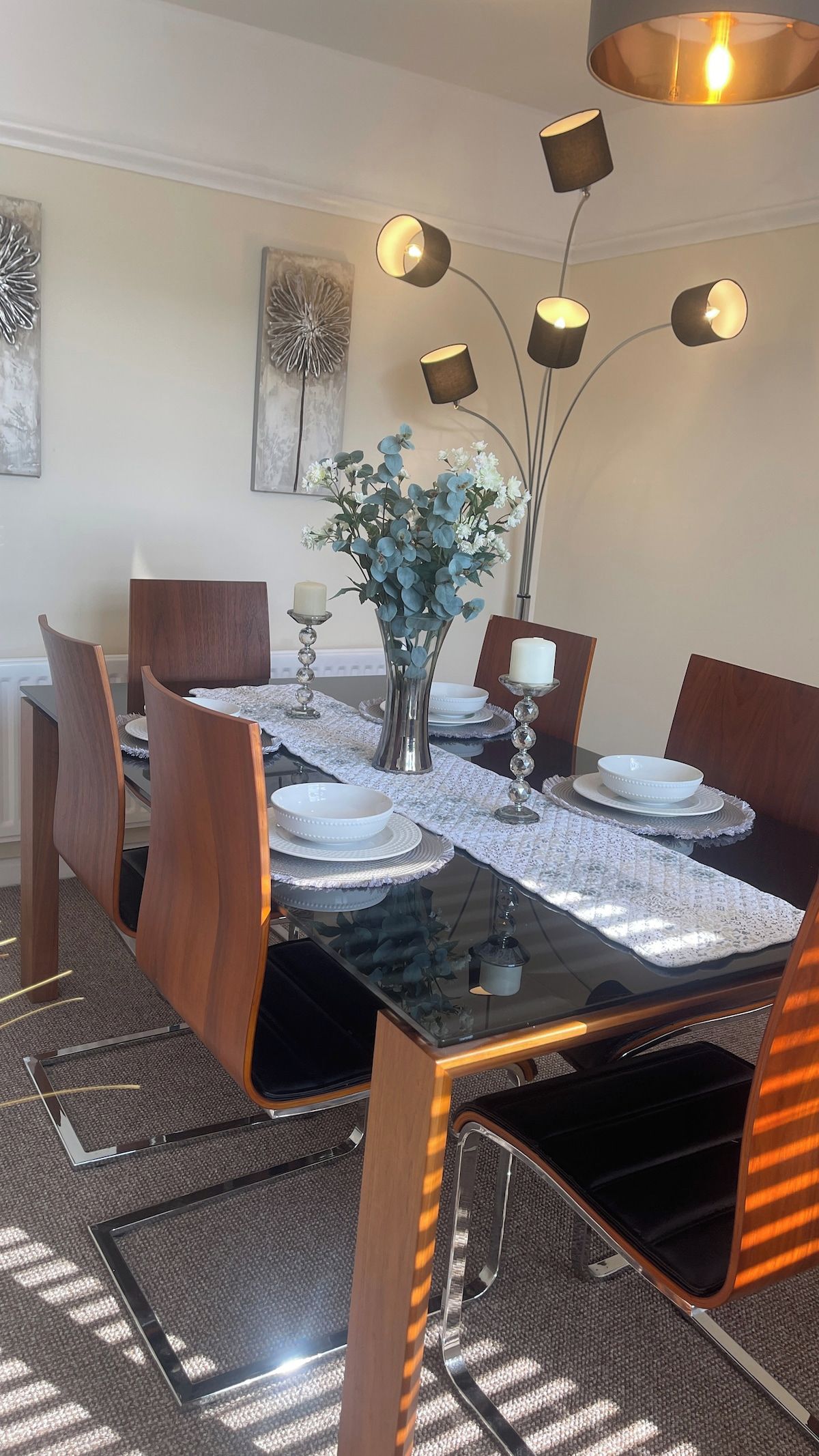 Ett matbord med en glasskiva, omgivet av trästolar, dukat med vita tallrikar och en vas med blå blommor.
