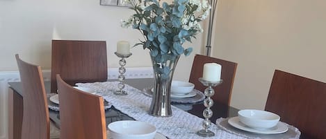 Une table à manger avec un plateau en verre, entourée de chaises en bois, dressée avec de l’assiette blanche et un vase rempli de fleurs bleues.