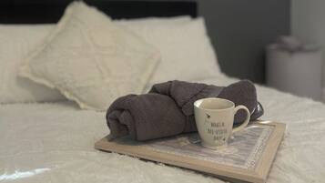 A neatly made bed with a rolled towel, a mug with a quote, and a framed picture on the bed.