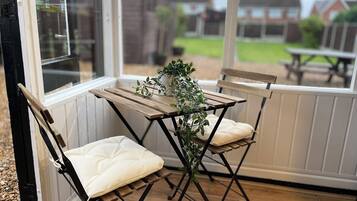 Eine Terrasse mit zwei Klappstühlen und einem Tisch, einem Topfpflanzen und Blick auf eine Wohngegend durch ein Fenster.
