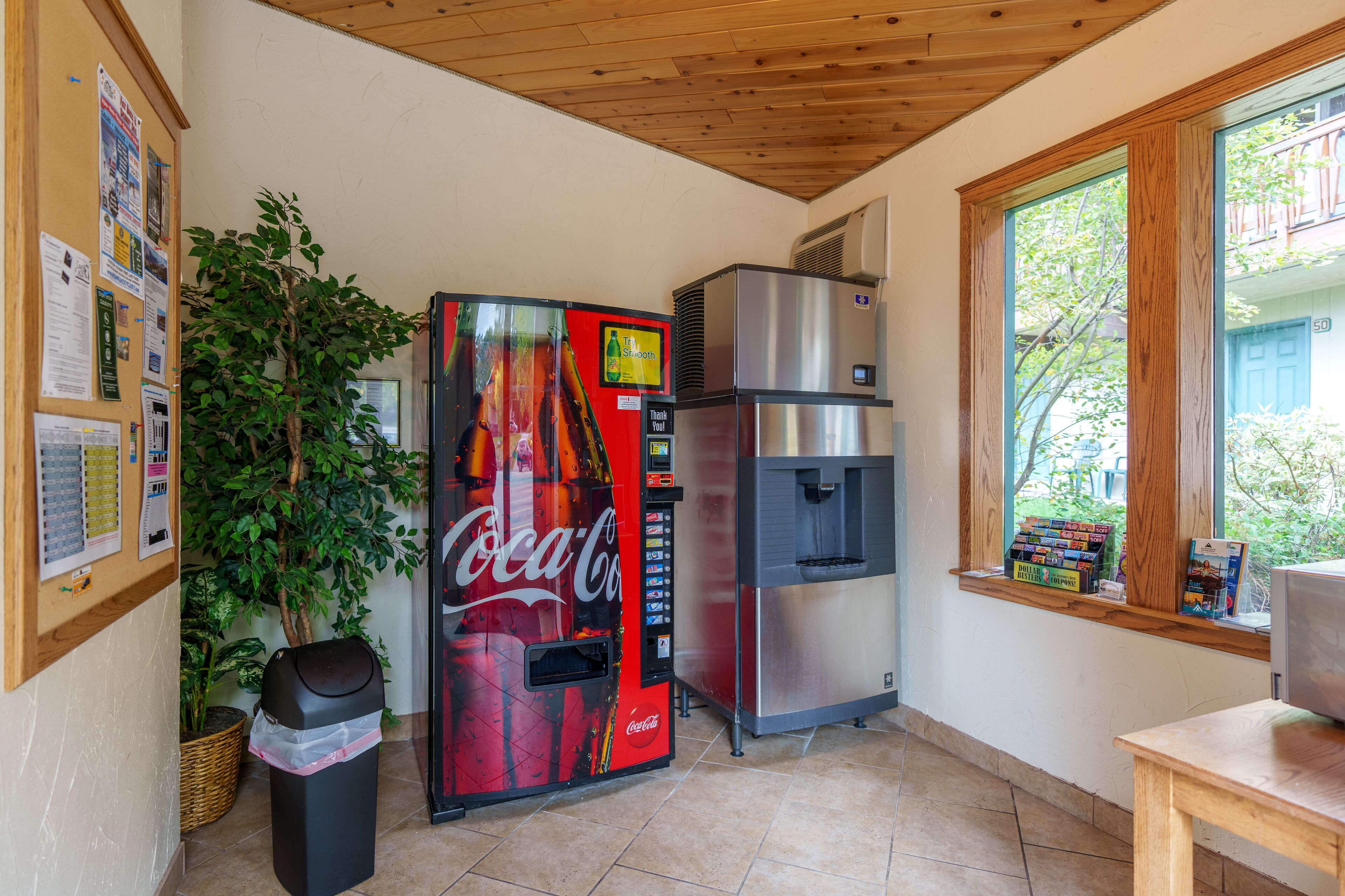 Cozinha com geladeira, bebedouro, máquina de Coca-Cola, lixeira e janela com vista para vegetação.