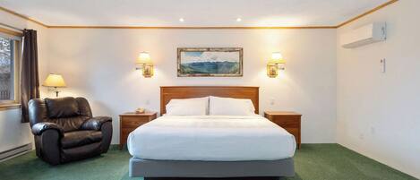 A hotel room with a large bed, a leather chair, two bedside tables, wall-mounted lamps, and a painting above the bed.