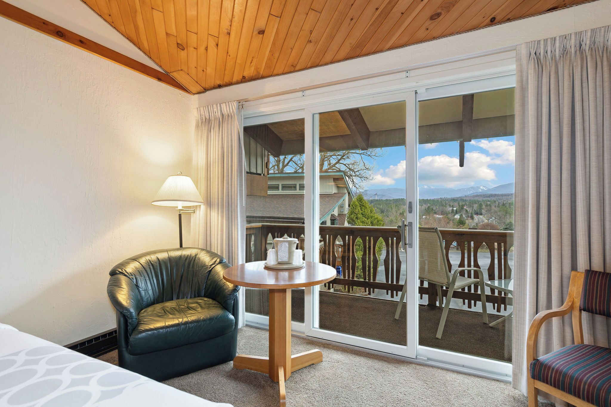 A hotel room with a wooden ceiling, a large sliding glass door leading to a balcony, a leather chair, a small round table with a lamp, and a bed with a patterned bedspread.