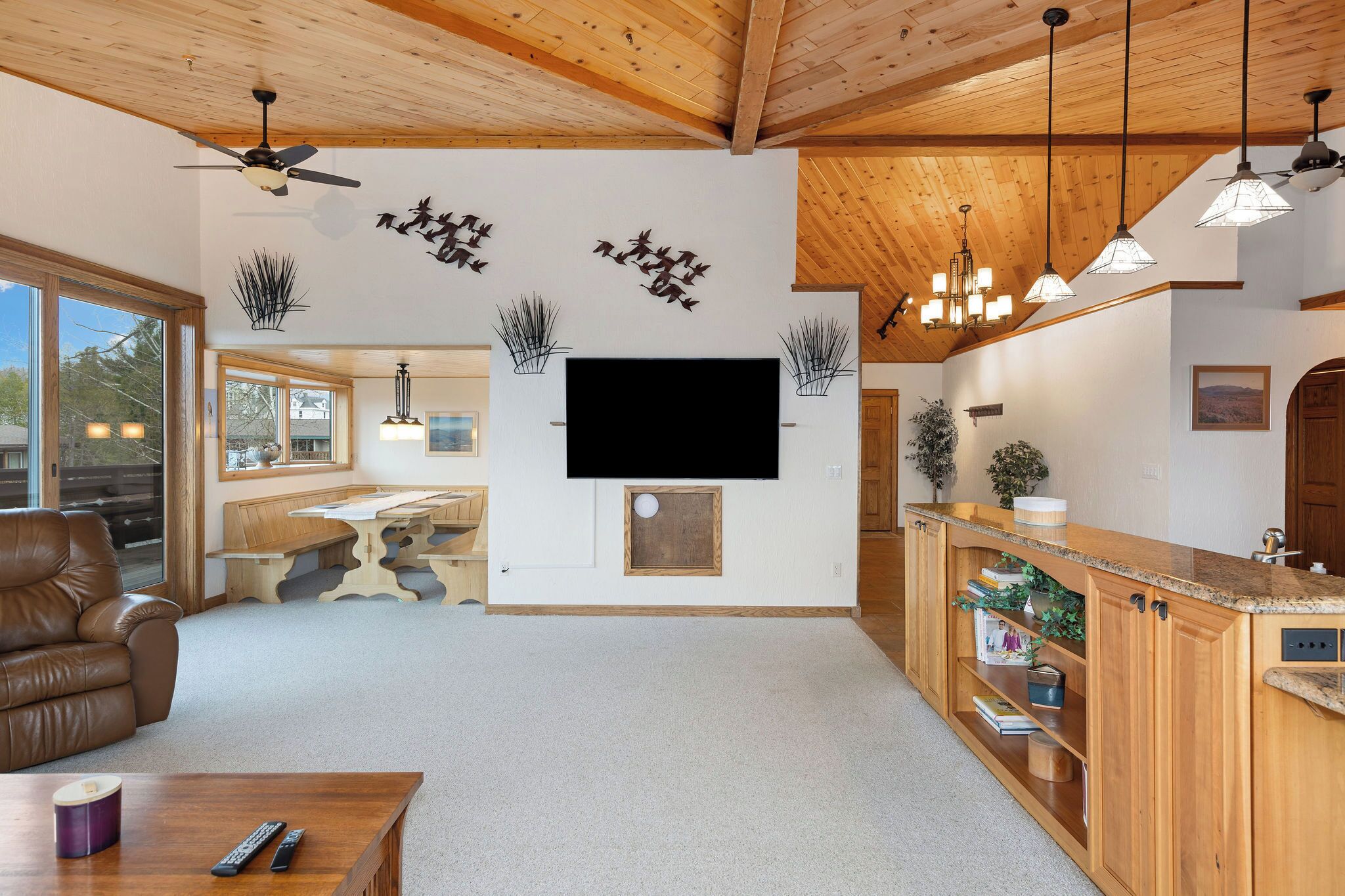 Ein geräumiges Wohnzimmer mit Holzboden, einem an der Wand montierten Flachbildfernseher, einem Ledersofa, einem Holztisch und einer Küchenzeile mit Holzausstattung und Pendelleuchten.