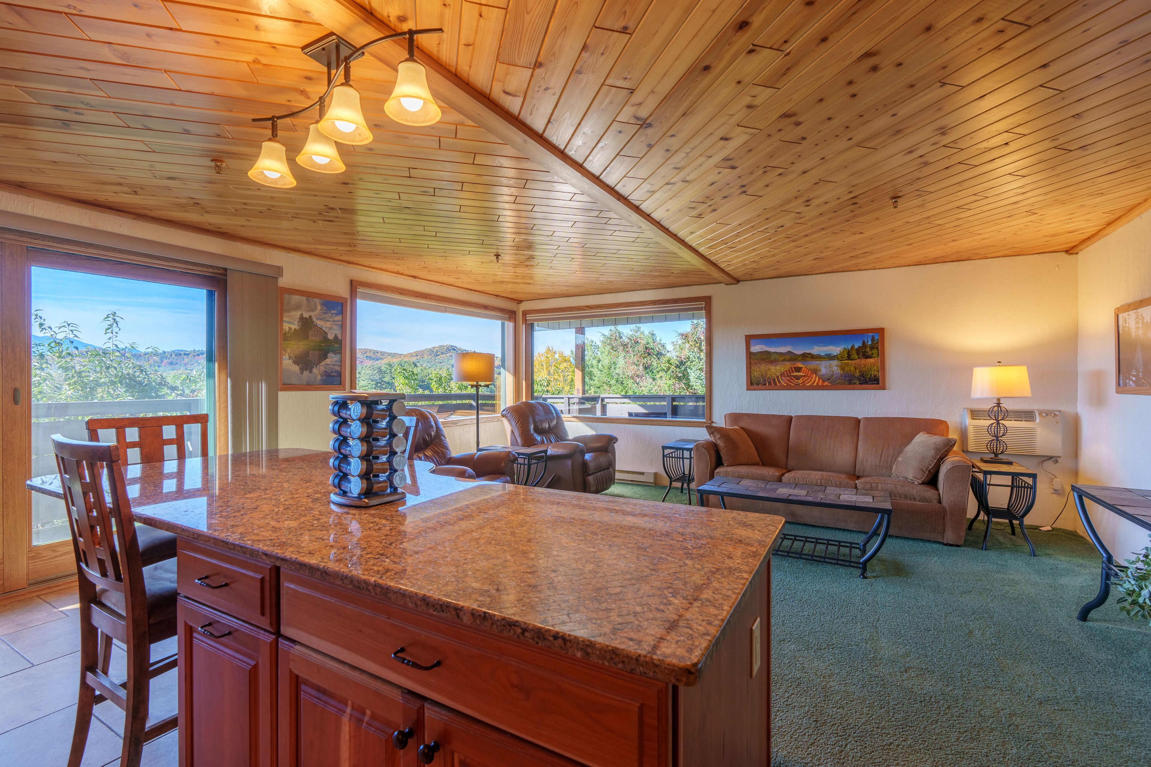A spacious living area with a wooden ceiling, a kitchen island, and large windows offering a view of the outdoors.