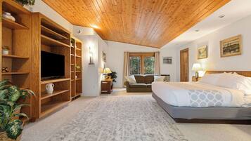 Une chambre avec un plafond en bois, un grand lit, un téléviseur posé sur une étagère en bois et une fenêtre avec des rideaux.
