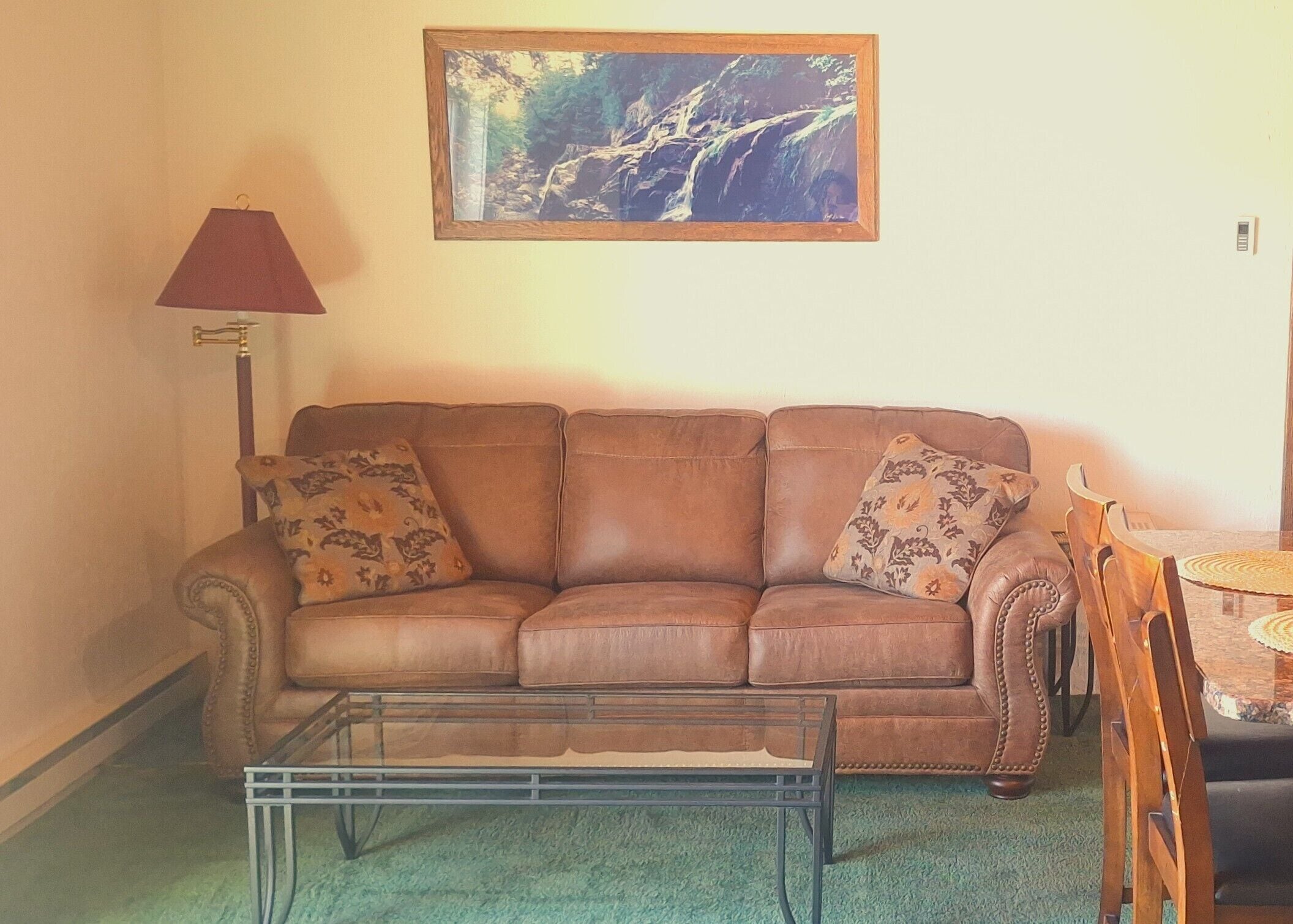 A brown leather sofa with patterned cushions, a glass coffee table, a red floor lamp, and a framed picture of a waterfall on the wall.