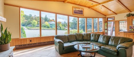 A spacious room with a green leather sectional sofa, a glass coffee table, and large windows offering a view of a lake and surrounding trees.
