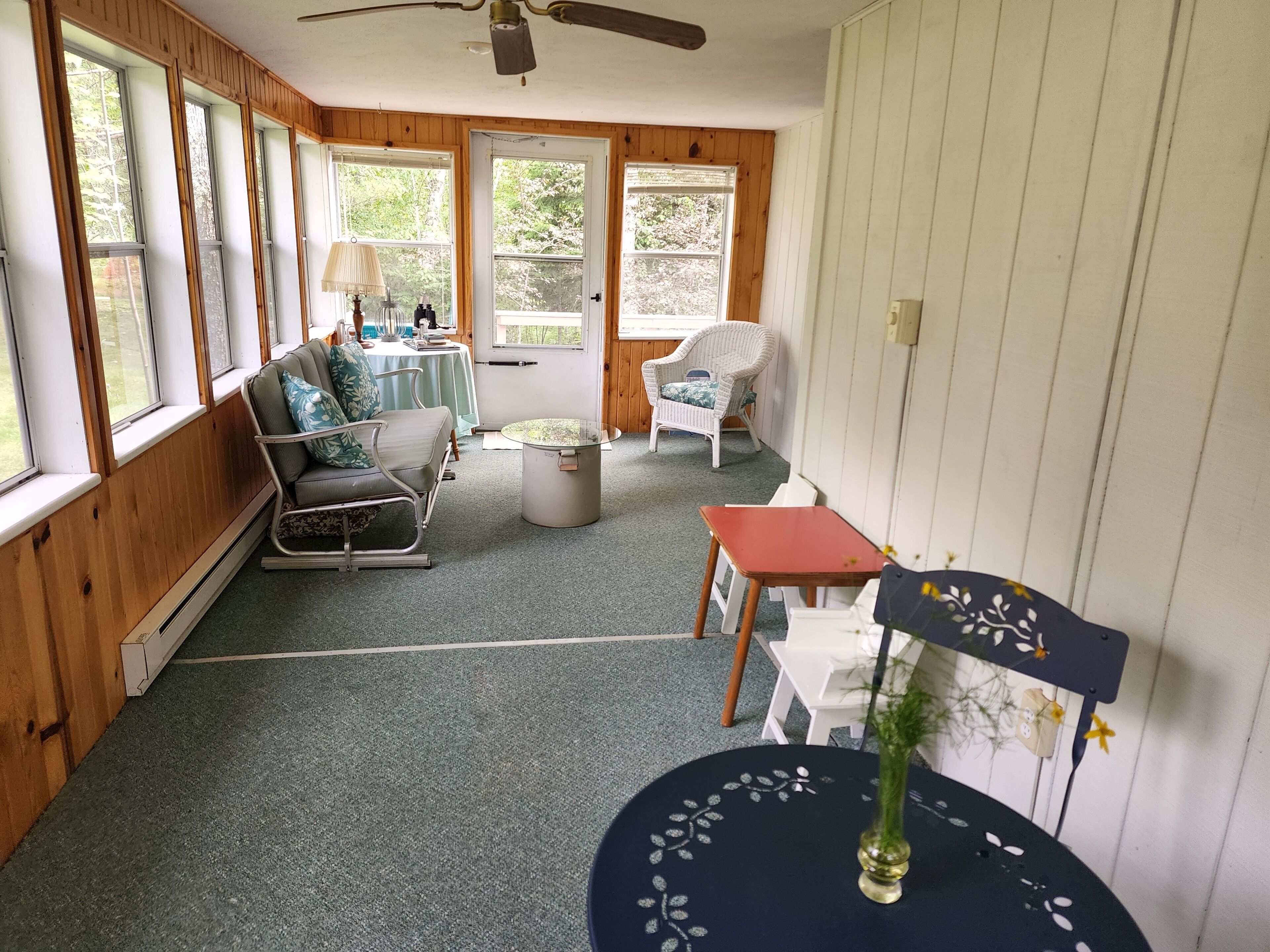 Una habitación acogedora con paredes de madera, un ventilador de techo, un sofá, una silla, una mesita y una ventana con vistas a árboles.