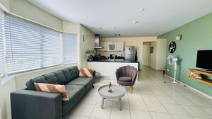 En moderne stue med en grå sofa, et rundt sofabord og en lænestol. I baggrunden er der et køkken med et køleskab og en komfur.