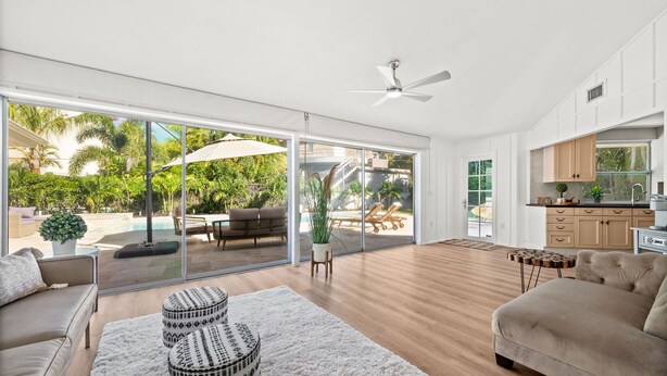 Un vaste espace de vie avec une grande porte-fenêtre coulissante donnant sur une piscine et un jardin, un ventilateur de plafond et un coin cuisine doté de meubles en bois.