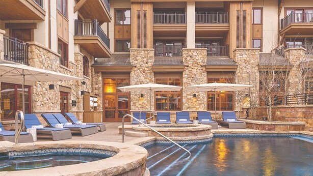 Un área de piscina con sillones y sombrillas, paredes de piedra y un edificio de varios niveles con balcones.