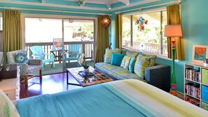 A room with a bed, a sofa, a coffee table, a bookshelf, and a balcony with chairs and a table. - Tracy's Tropical Treasure #2 earned "Best in Kihei" four times and “#1 in Maui” (Kihei)