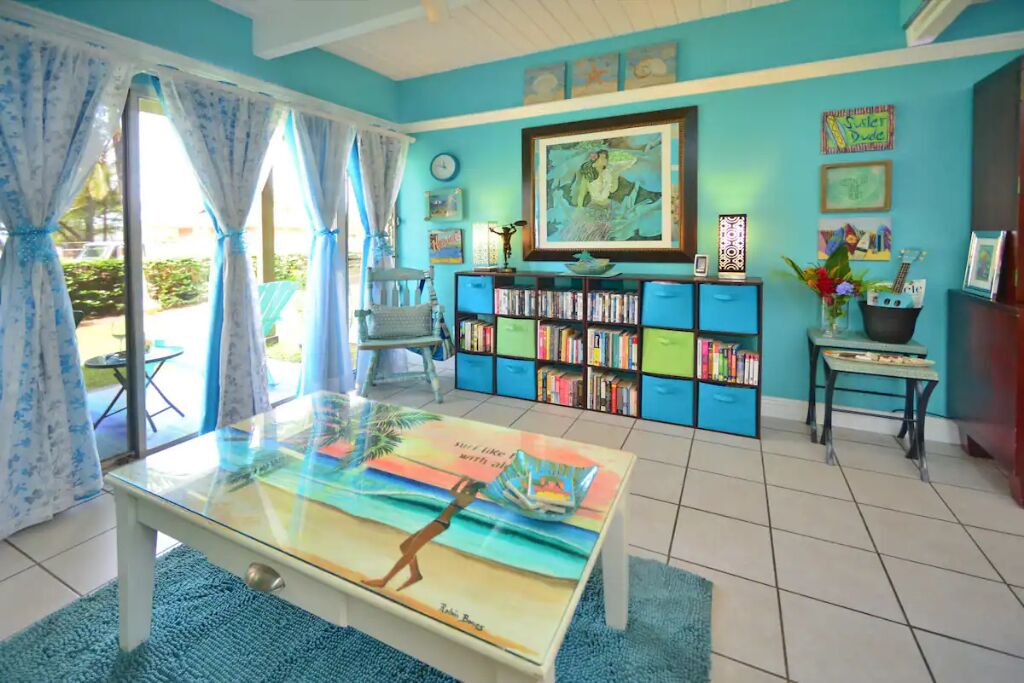 Een kamer met een blauwe muur, een boekenkast, een glazen salontafel met een strandmotief en een stoel.