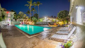 A swimming pool with lounge chairs and palm trees, surrounded by buildings. - Tracy's Tropical Treasure #3 earned "Best in Kihei" four times and “#1 in Maui” (Kihei)