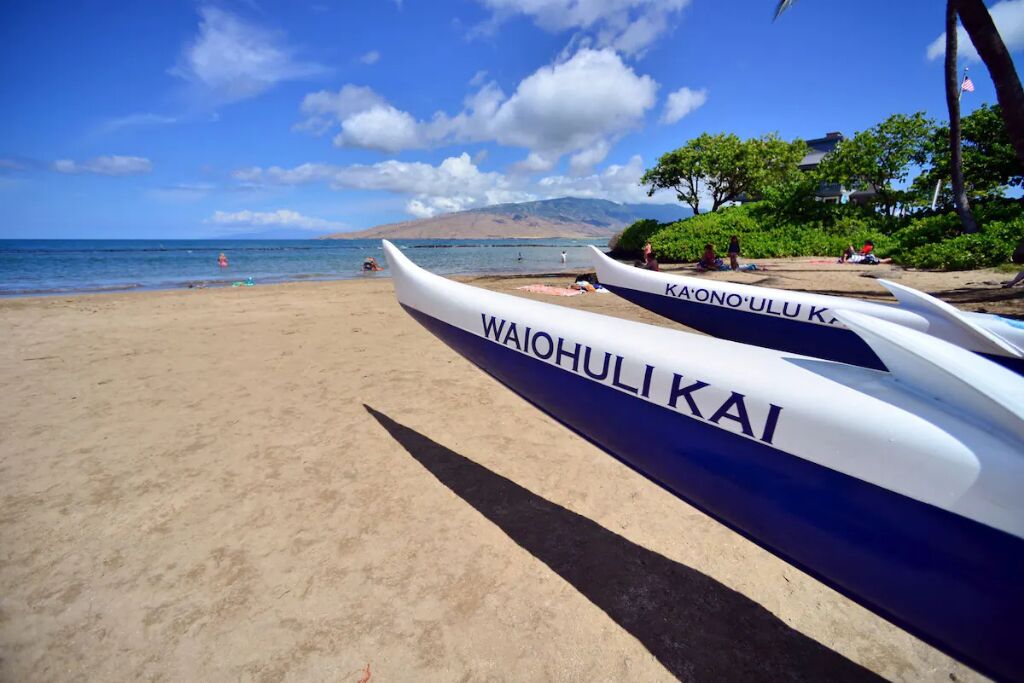 Una playa con canoas identificadas con los rótulos 'WAIOHULI KAI' y 'KA'ONO'ULU KAI' sobre la arena.