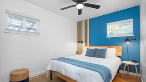 A bedroom with a bed, a ceiling fan, a bedside table, a lamp, and a window with blinds.