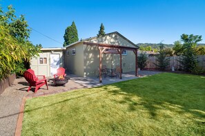 Ein Garten mit einer überdachten Terrasse, roten Adirondack-Stühlen, einer Feuerstelle und einem gepflegten Rasen.