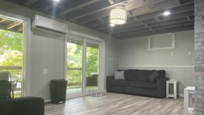 Een moderne woonkamer met een grijze bank, een klein witte tafel en een tv-meubel. Er is een schuifdeur die leidt naar een buitenruimte met een patio stoel.