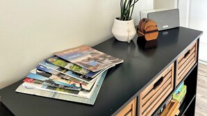 Una habitación con un mueble bajo negro para el televisor, una planta blanca en maceta, un adorno de madera y una pila de revistas.