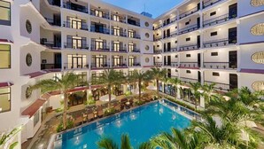 A multi-story hotel with a central courtyard featuring a swimming pool, palm trees, and outdoor seating areas.